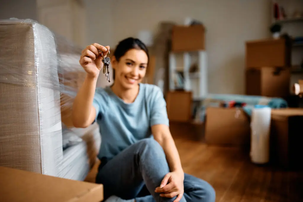 Smiling woman holding up the keys to her first home among moving boxes
